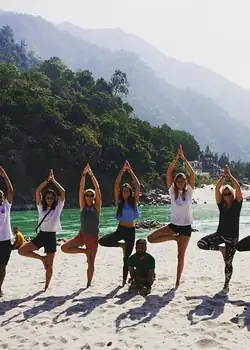 Students in tree pose on a river beach with forested hills behind
